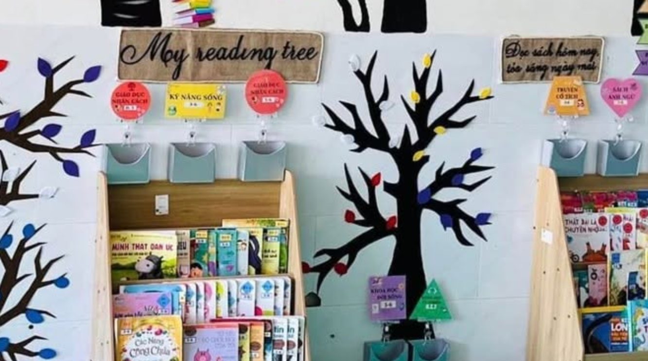 Góc thư viện lớp mầm non với hai kệ sách Montessori đặt dưới tranh cây đọc sách